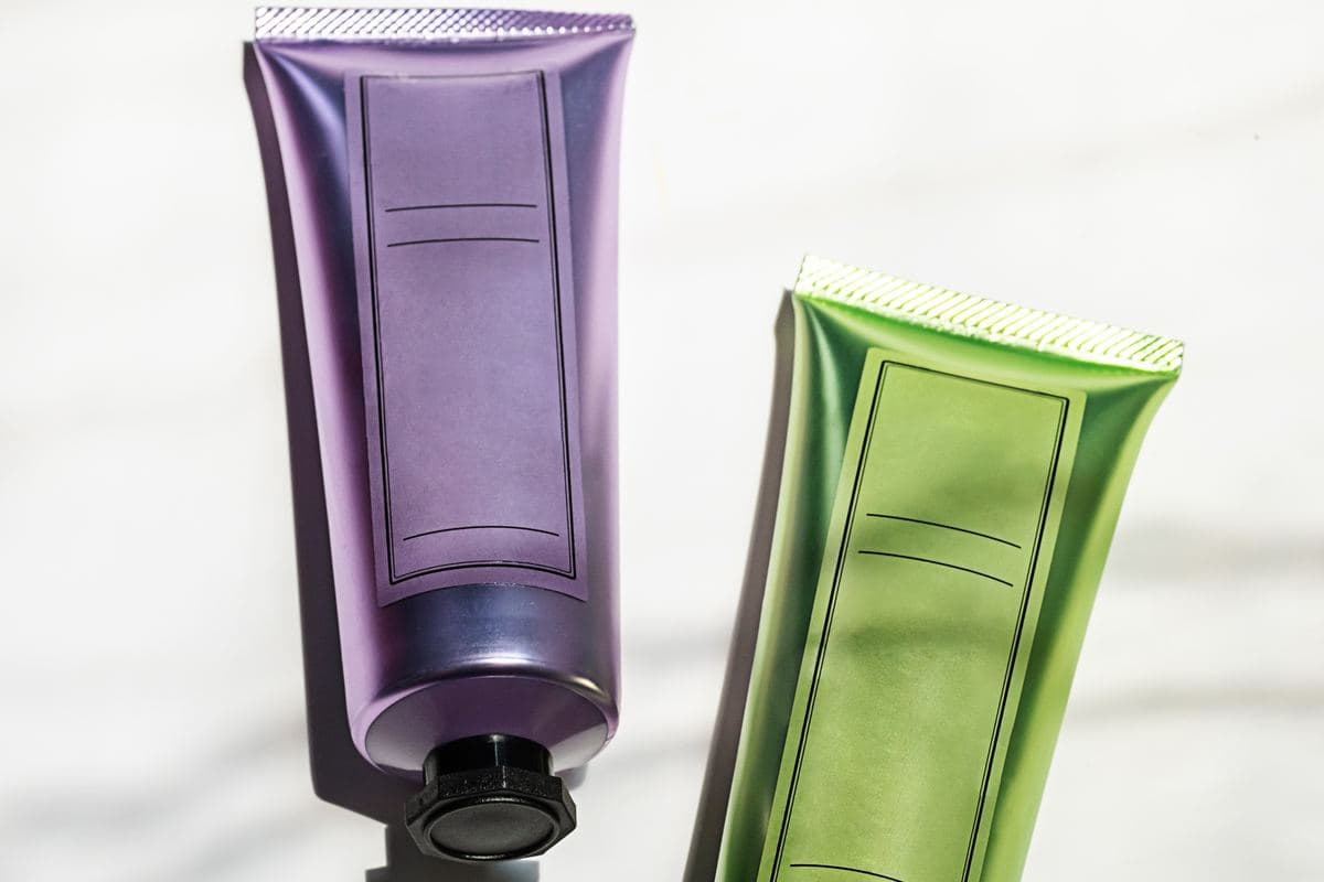 Dos tubos de cosmética sin etiqueta, uno morado y otro verde, sobre mármol blanco con sombras marcadas