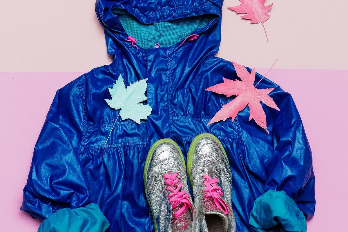Un chubasquero azul brillante extendido en cenital sobre fondo rosa, con unas zapatillas plateadas y hojas pintadas de rosa